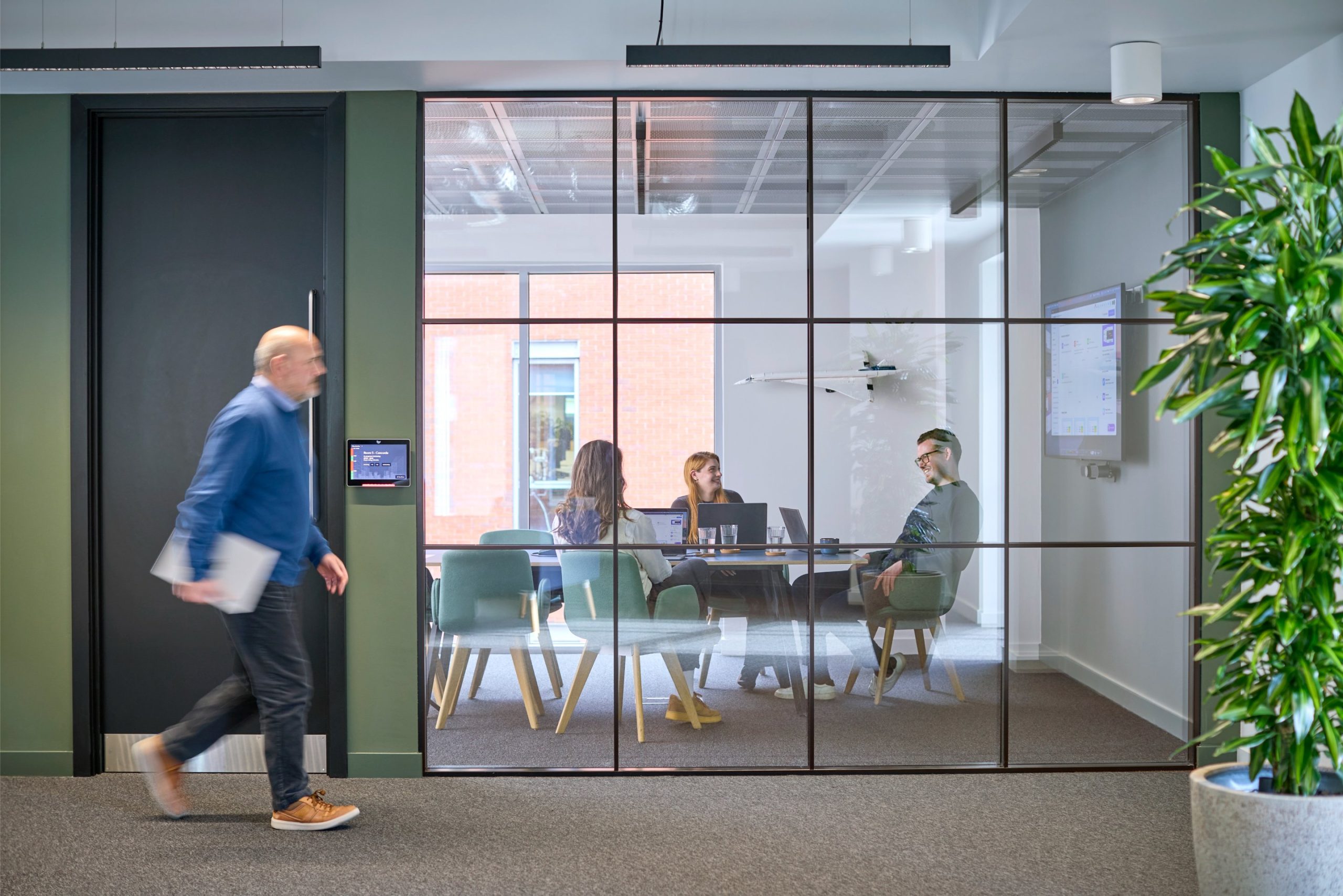A man walks past a glass-walled meeting room where four people are engaged in a discussion around a table with laptops and drinks.