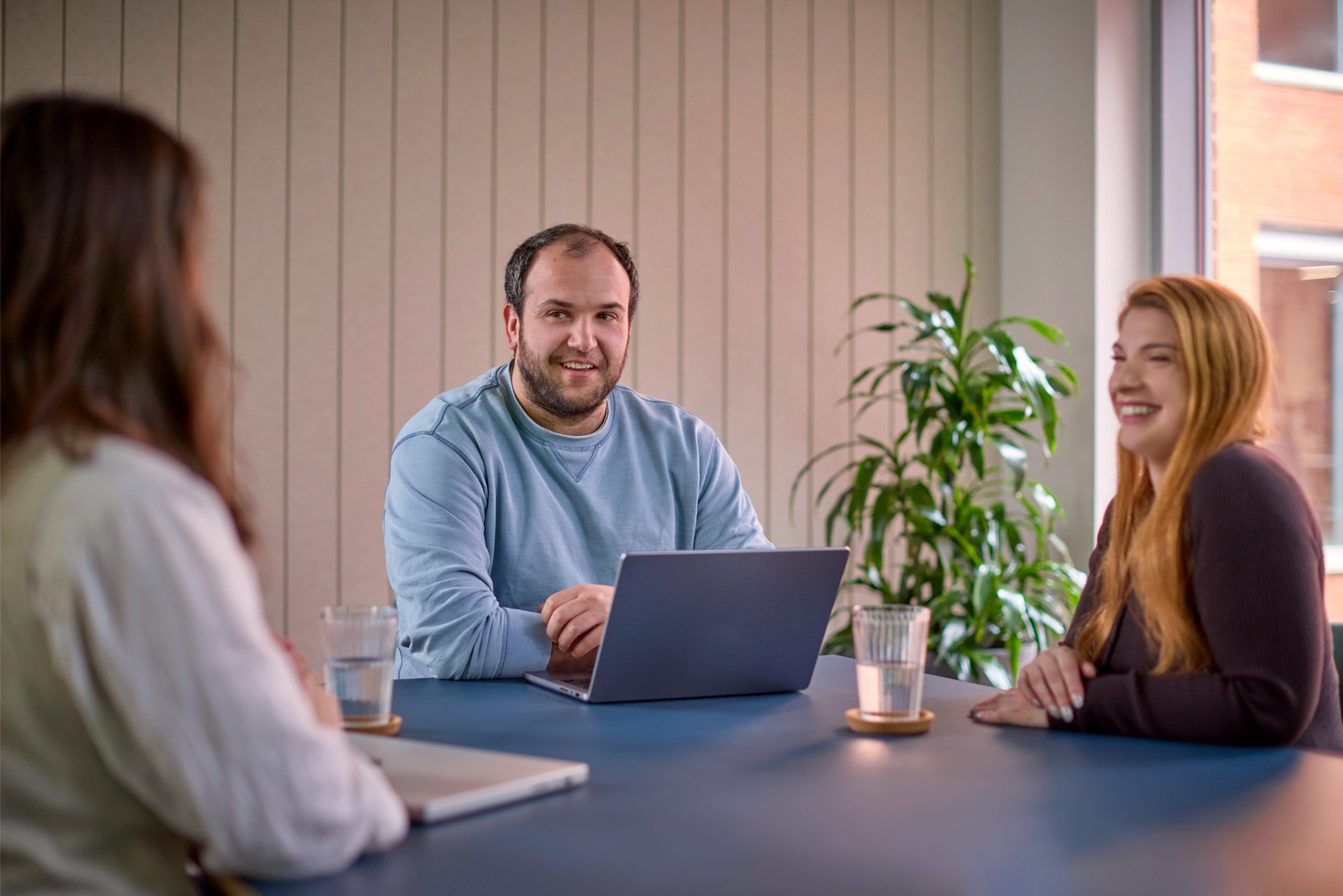 Three people engaged in a discussion around a table, with a laptop and glasses of water in a modern office setting.