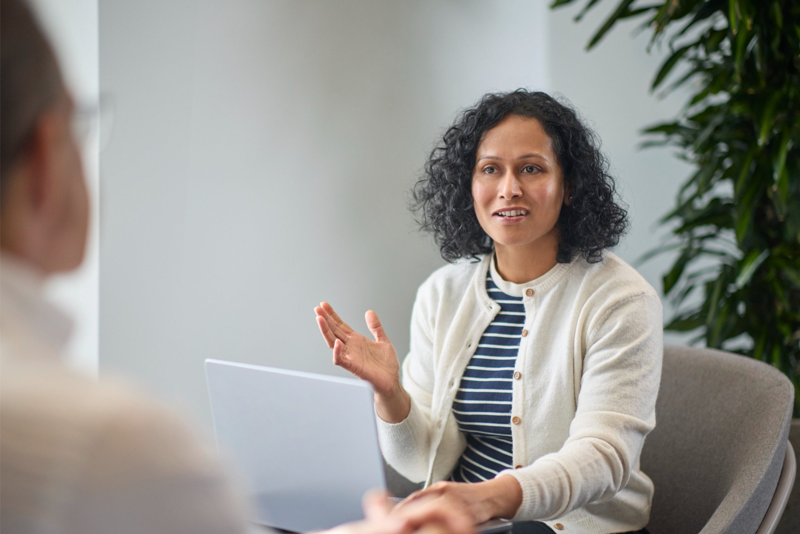A woman with curly hair wearing a striped shirt and cardigan gestures while speaking during a conversation in an office setting.