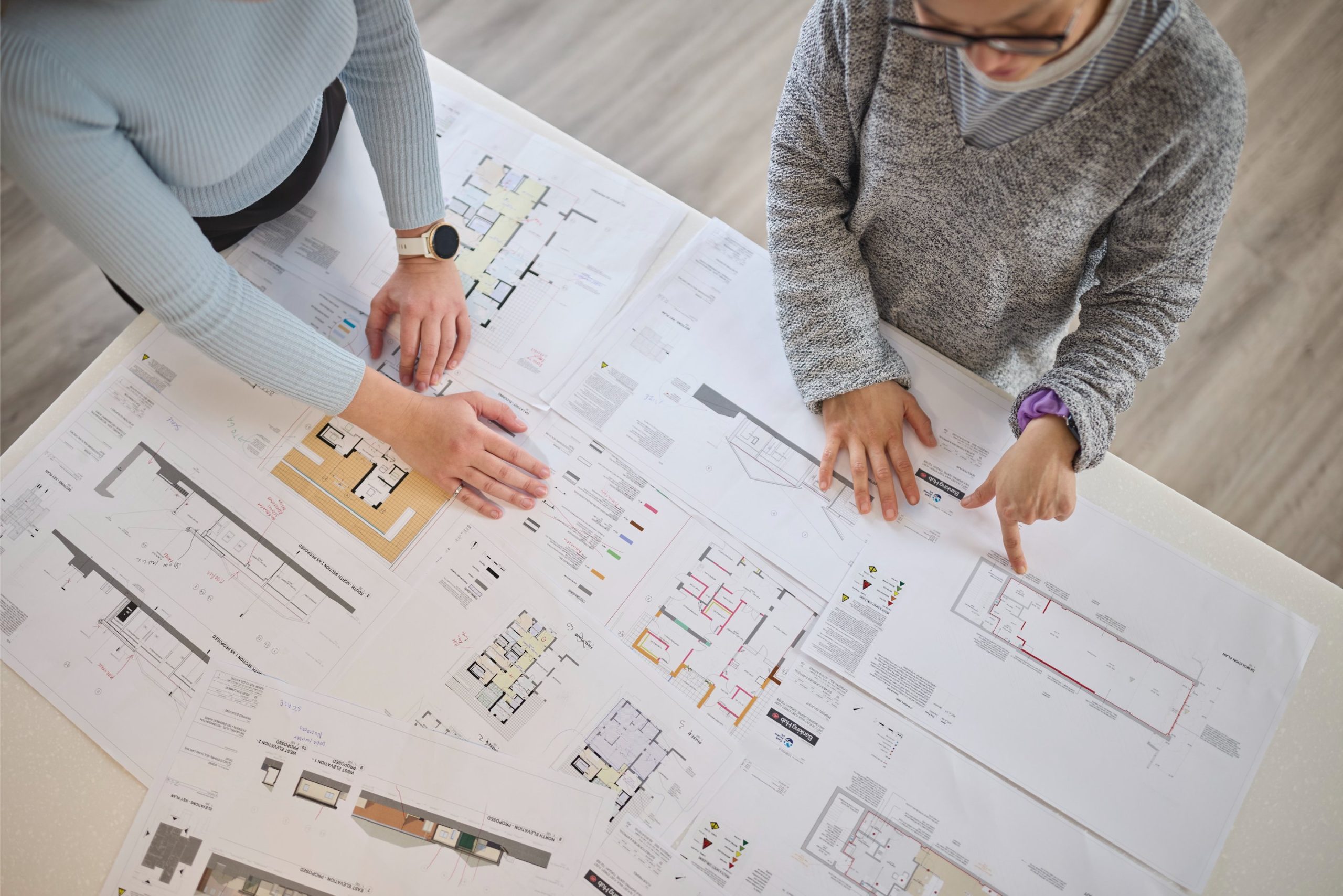 Two people examining architectural plans on a table, with various floor layouts and notes visible.