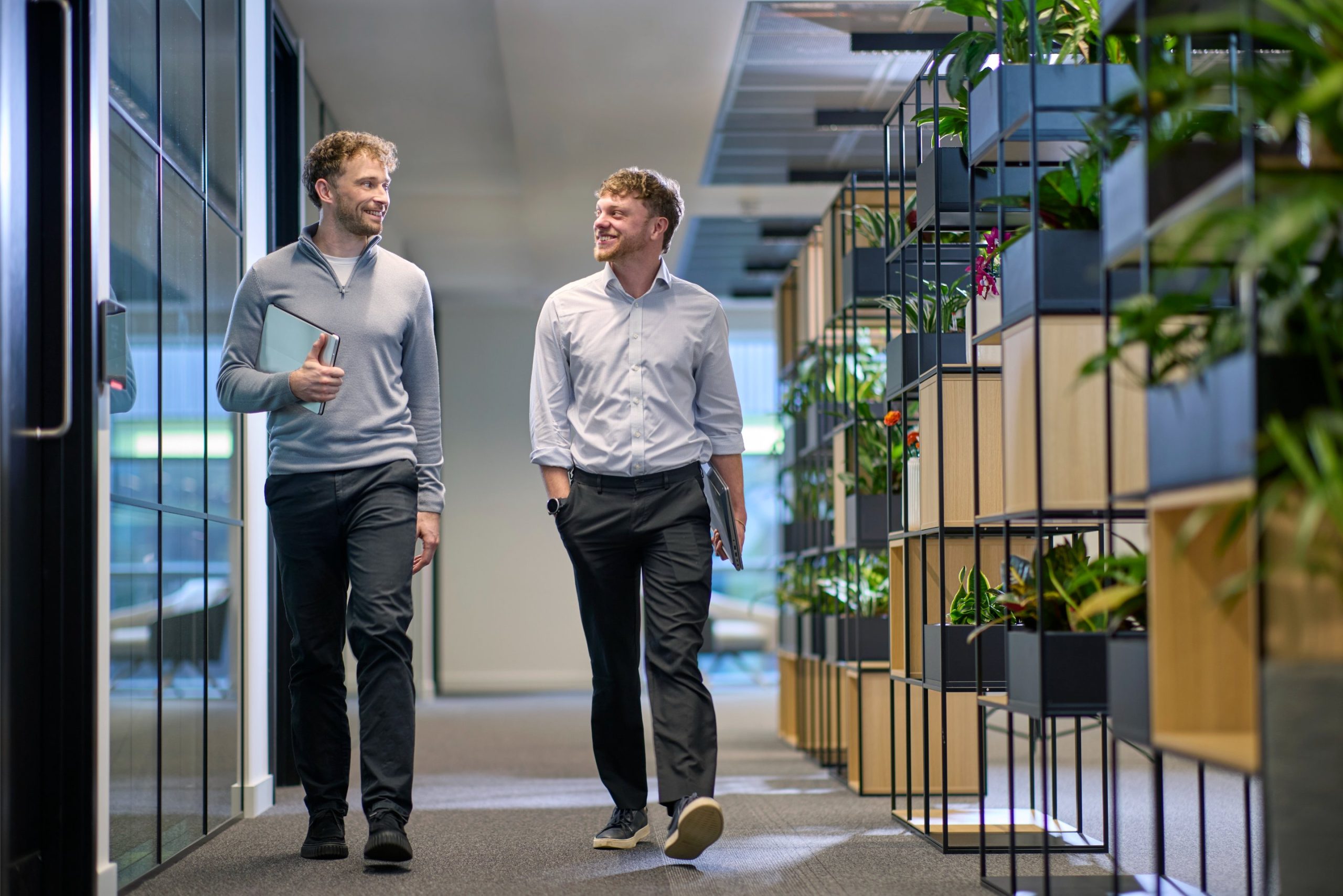 Two men walking and smiling in a modern office corridor with greenery and planters along the walls.