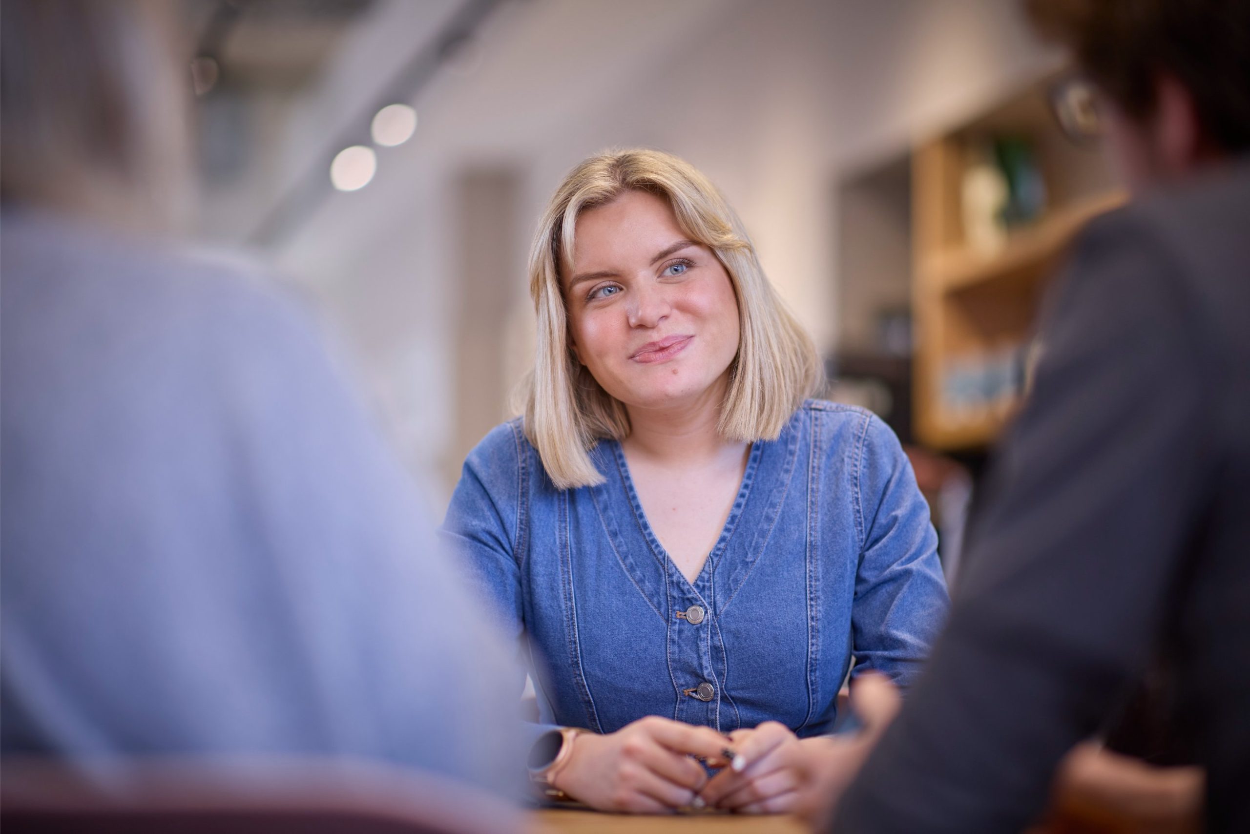 A woman with shoulder-length blonde hair, wearing a denim shirt, sits at a table engaged in conversation with two out-of-focus individuals.