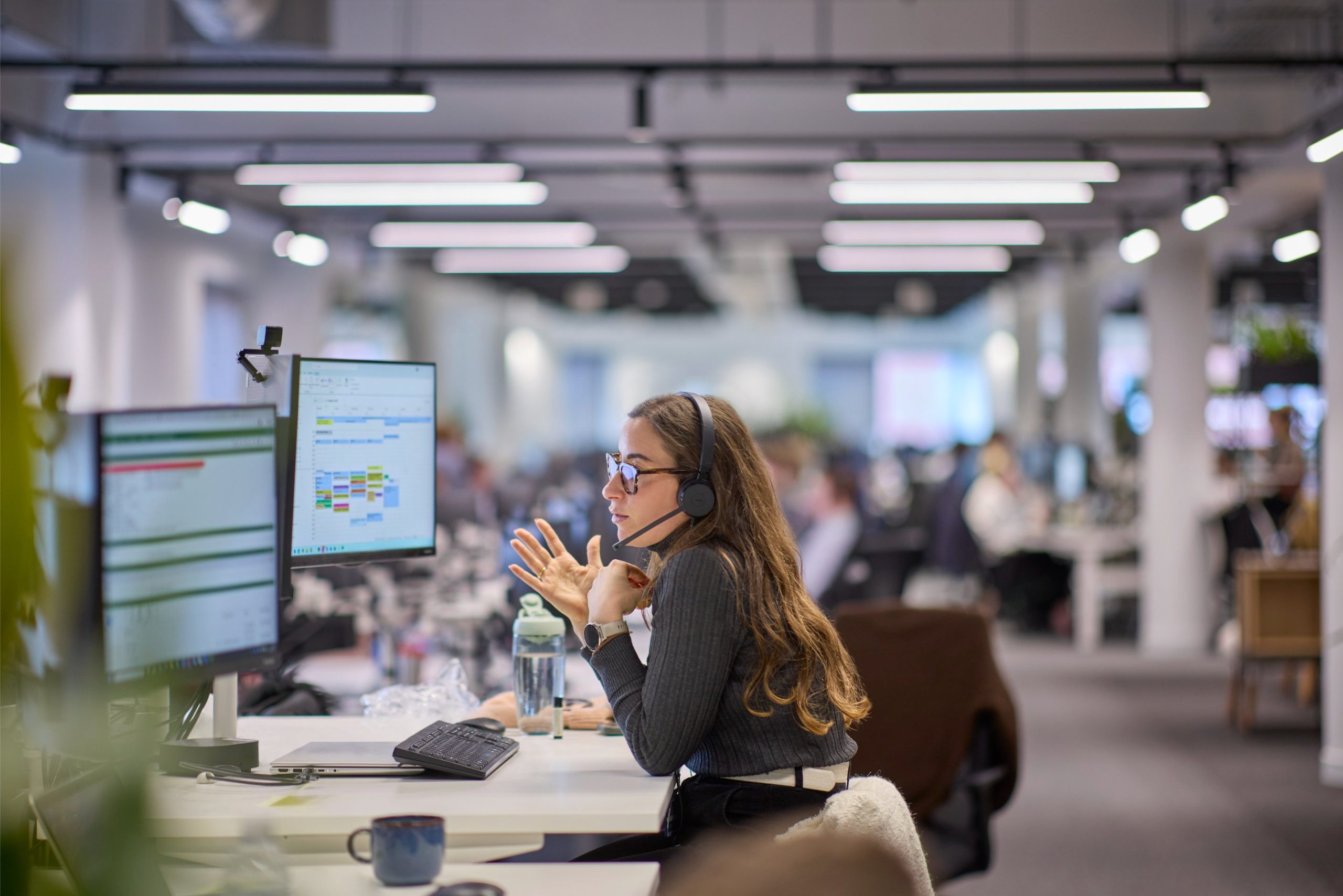 A woman wearing a headset gestures while speaking at a desk with multiple computer monitors in a busy office environment.