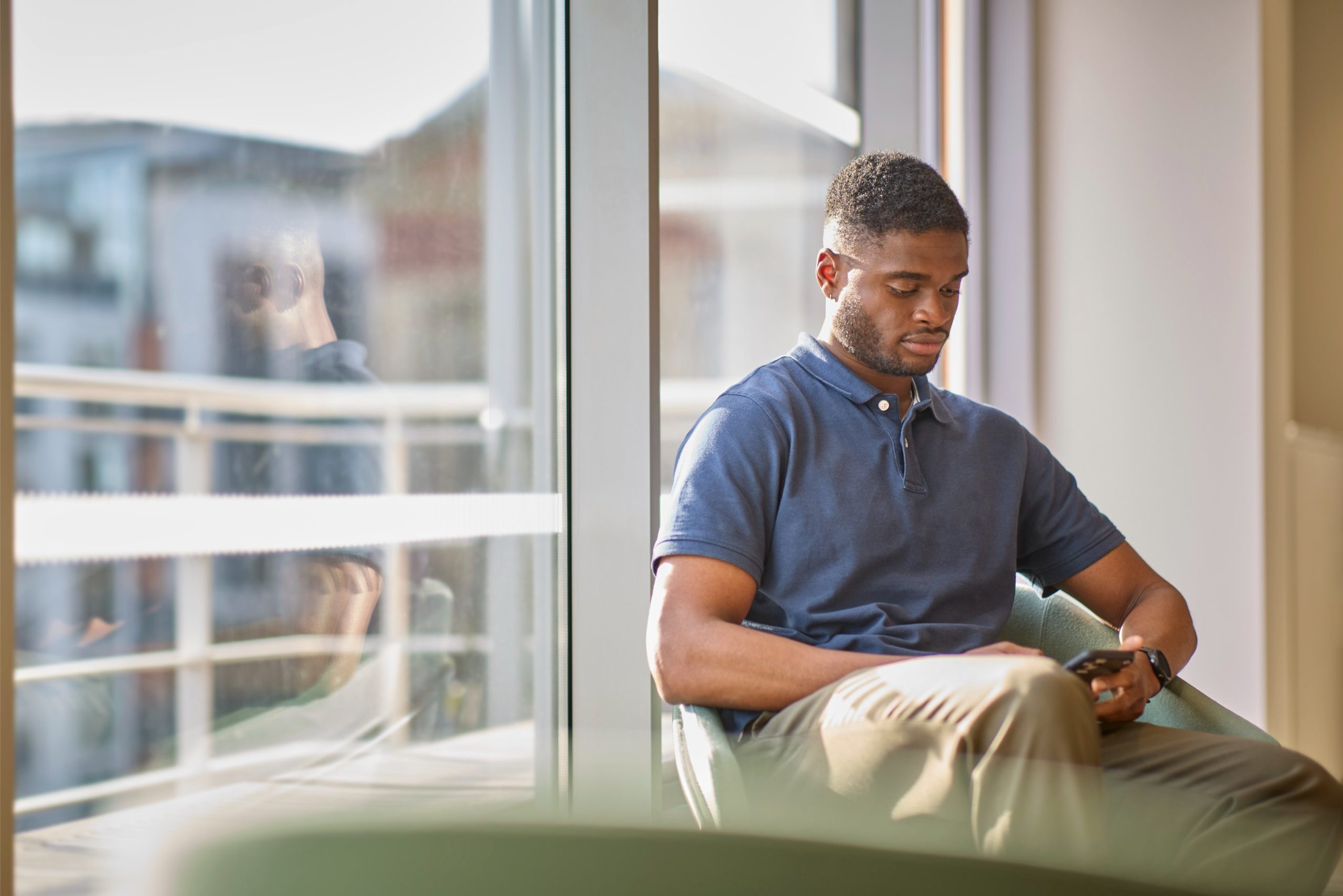 A young man sitting in a chair, focused on his smartphone, with large windows and a blurred outdoor view in the background.