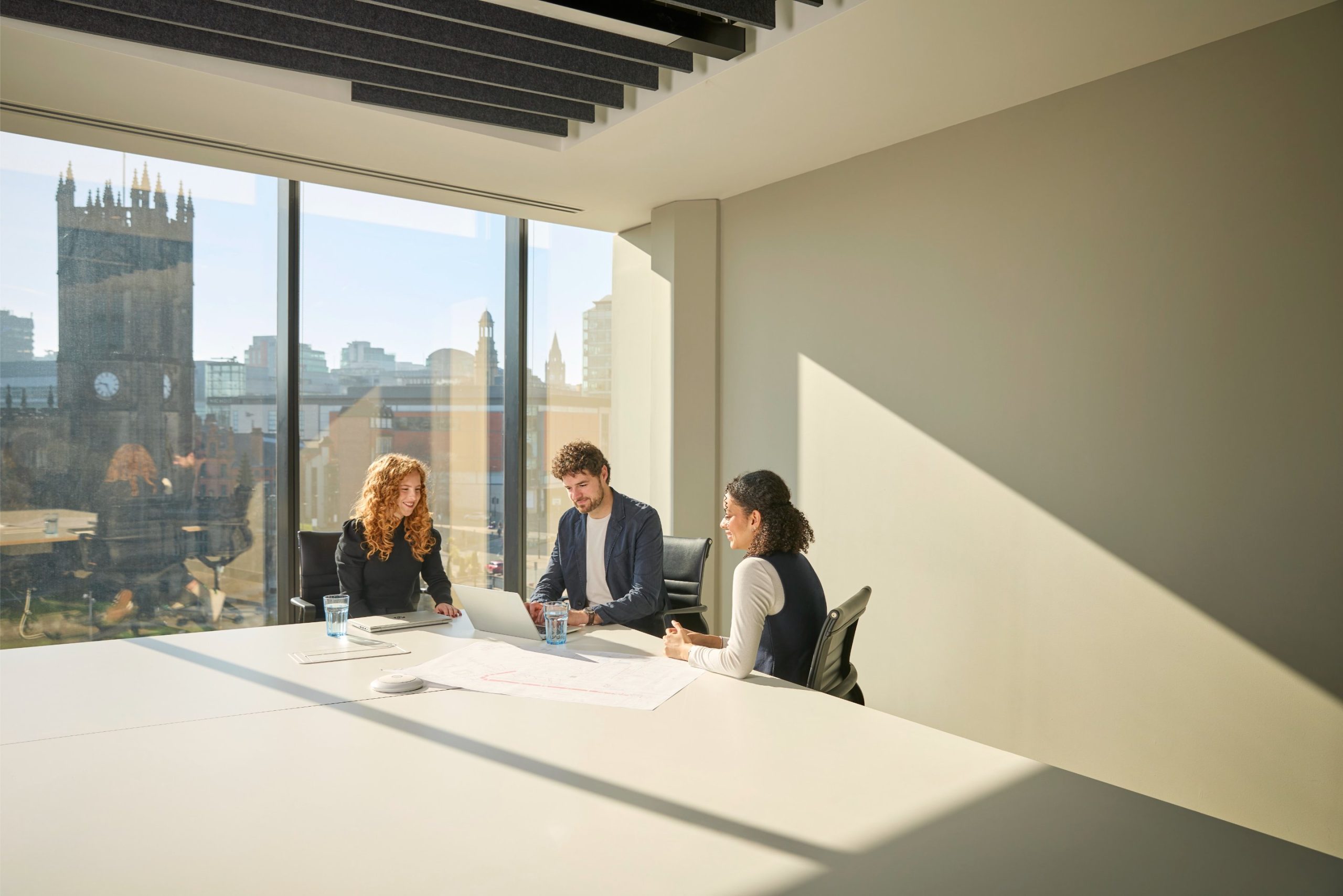 Three professionals collaborate at a conference table with a view of a cityscape, including a clock tower, in a well-lit office space.