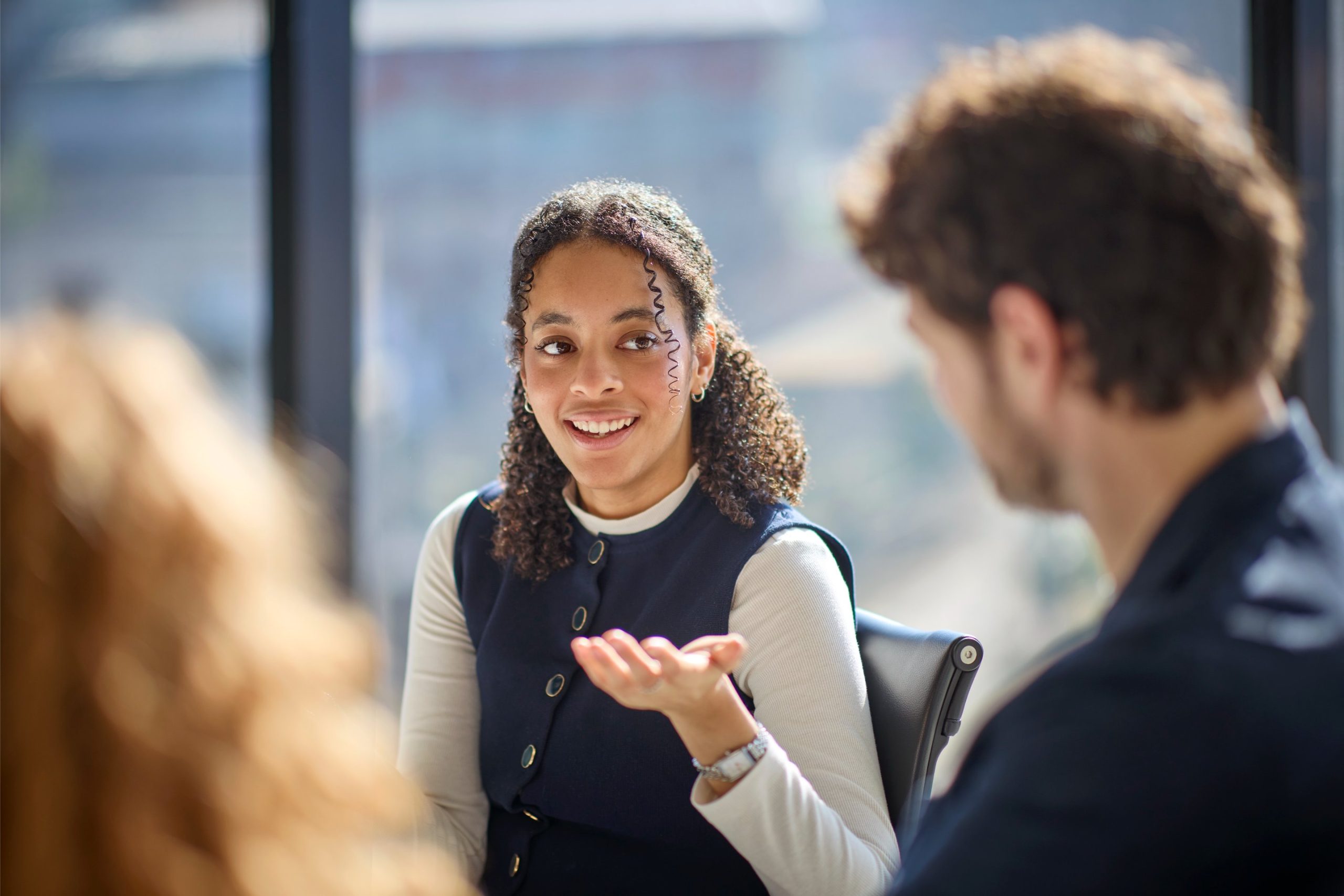 A woman with curly hair is engaged in conversation, gesturing with her hand while seated in a modern office setting.