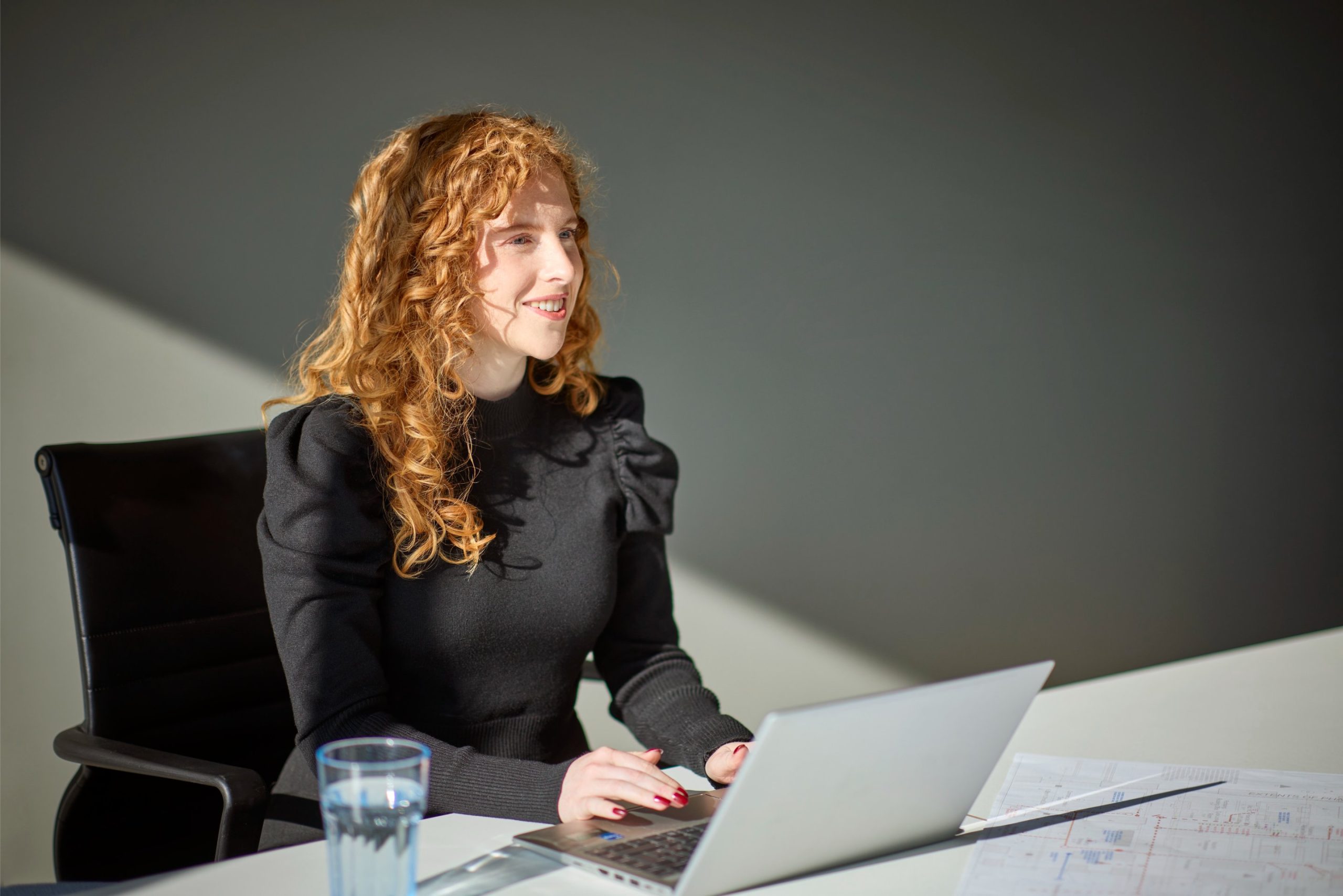 A woman with curly red hair smiles while working on a laptop at a desk, with a glass of water and documents visible nearby.