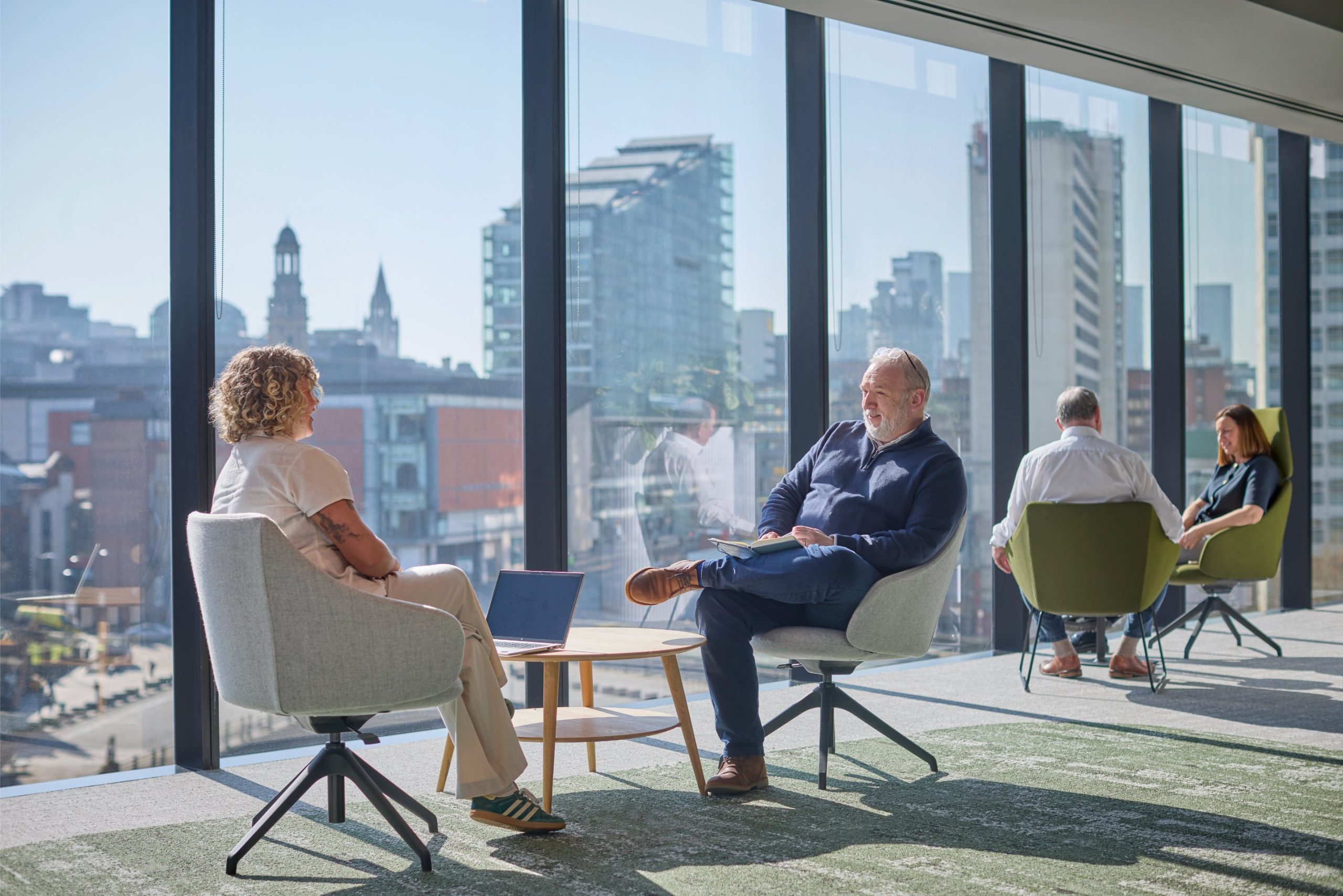 Two people engage in conversation in a modern office space with large windows overlooking a cityscape, while two others are seated in the background.