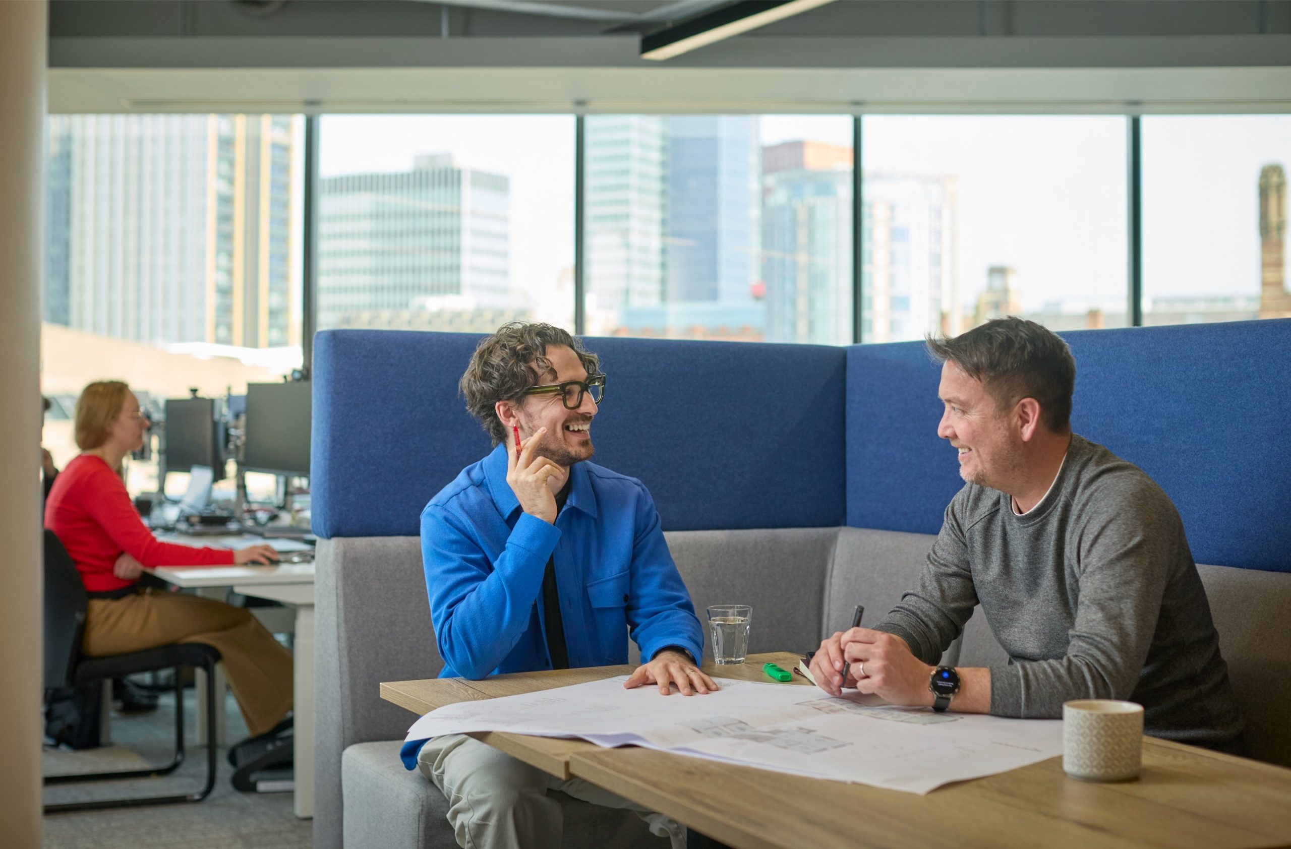Two men engaged in a discussion at a table with architectural plans, while a woman works at a computer in the background.