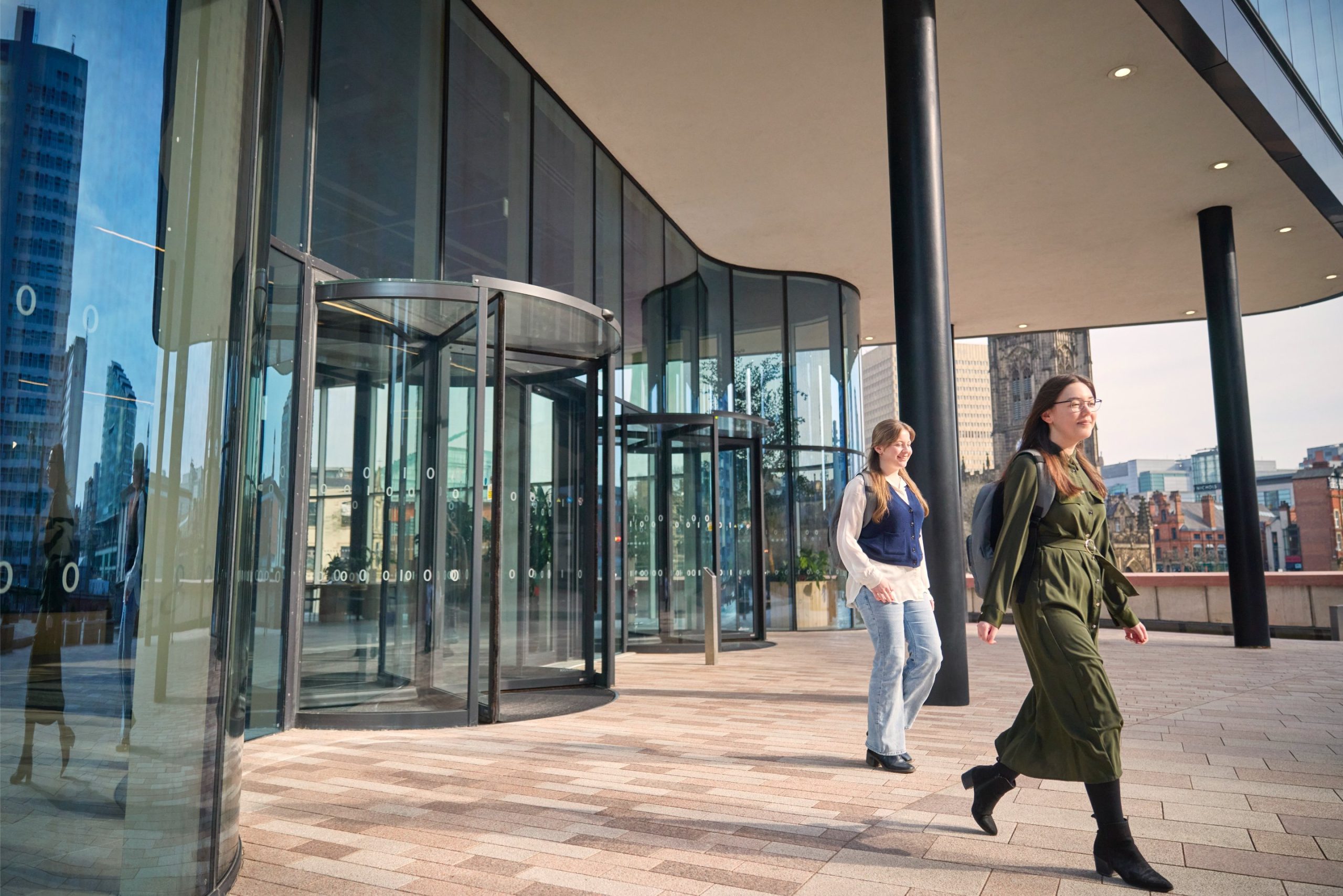 Two women walking outside a modern glass building with a revolving door, reflecting cityscape elements in the glass.