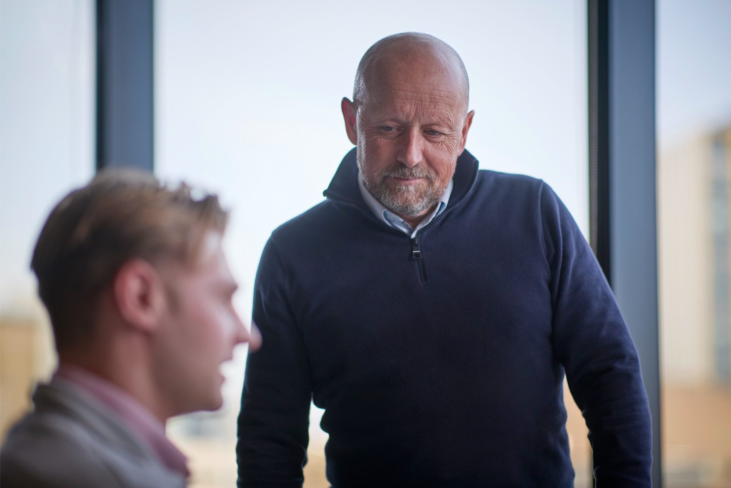 Two men are engaged in conversation, with one man looking thoughtfully at the other, who is partially turned away. The setting appears to be an office with larg