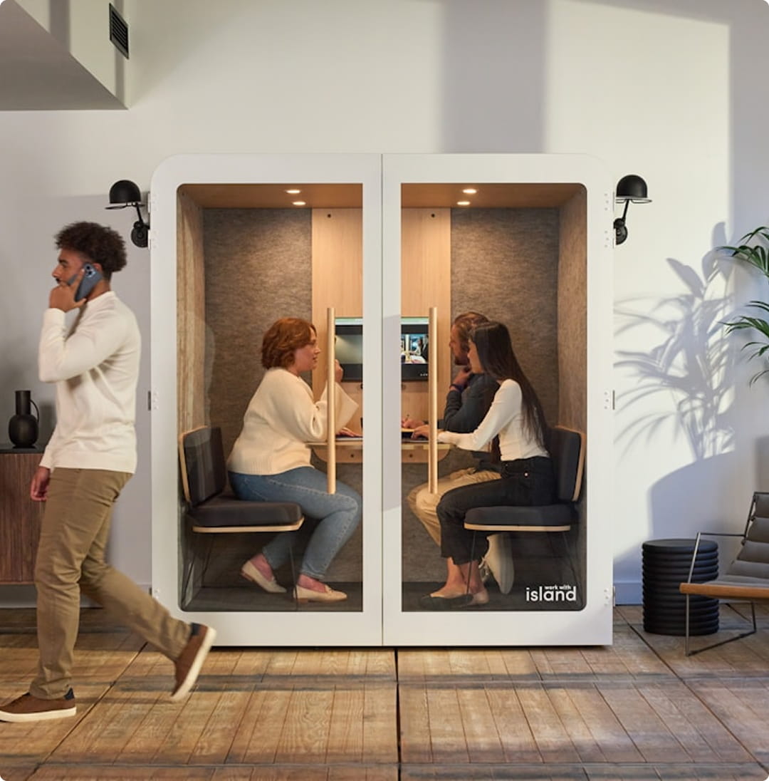 A man walks past a soundproof booth where two people are working at computers, while another person sits inside the booth talking to them.